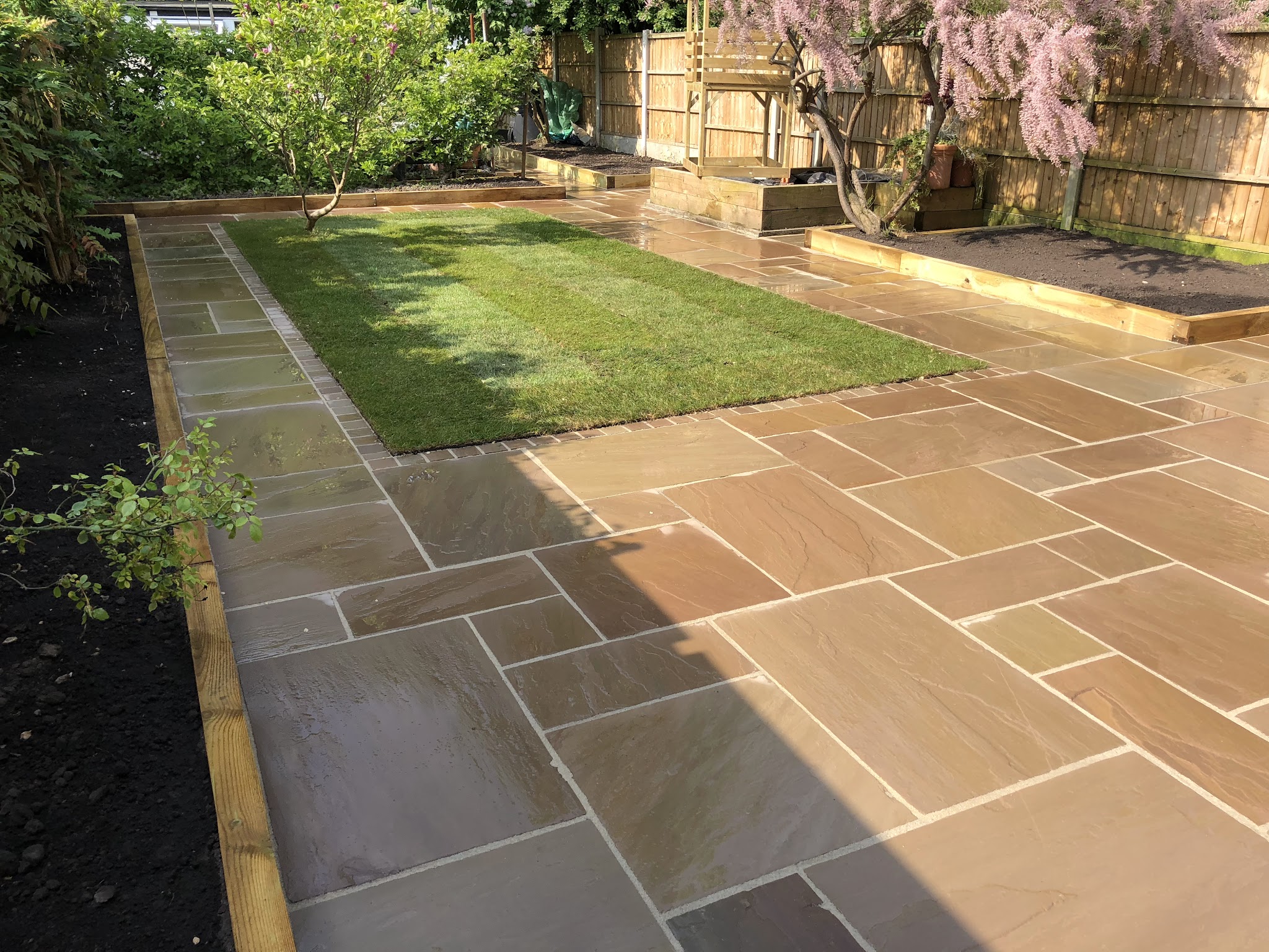 A buff riven patio wrapping round a lawn with sleeper-edged beds