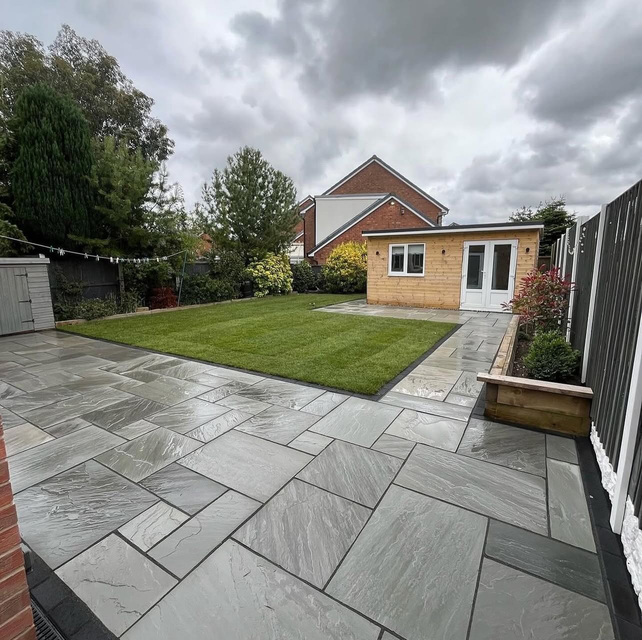 A grey riven patio, lawn and summer house under a wide sky